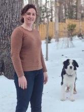 A white woman with curly hair stands in the snow wearing a brown sweater and jeans. There is a black and white border collie behind her.