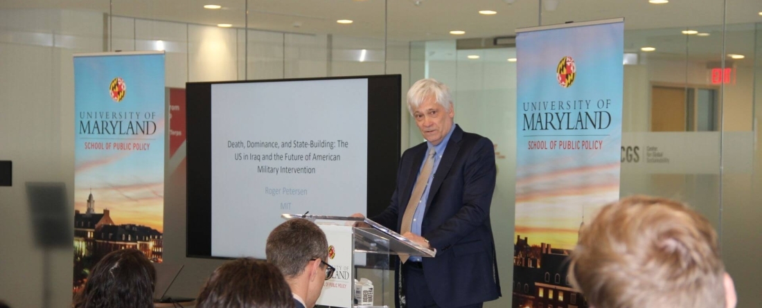 Roger Petersen stands against a glass wall with a TV behind him stating the name of his book, "Death, Dominance, and State-Building" and an SPP banner on each side of him